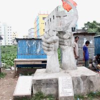 a concrete monument of a hand with people standing nearby - Rana Plaza Monument