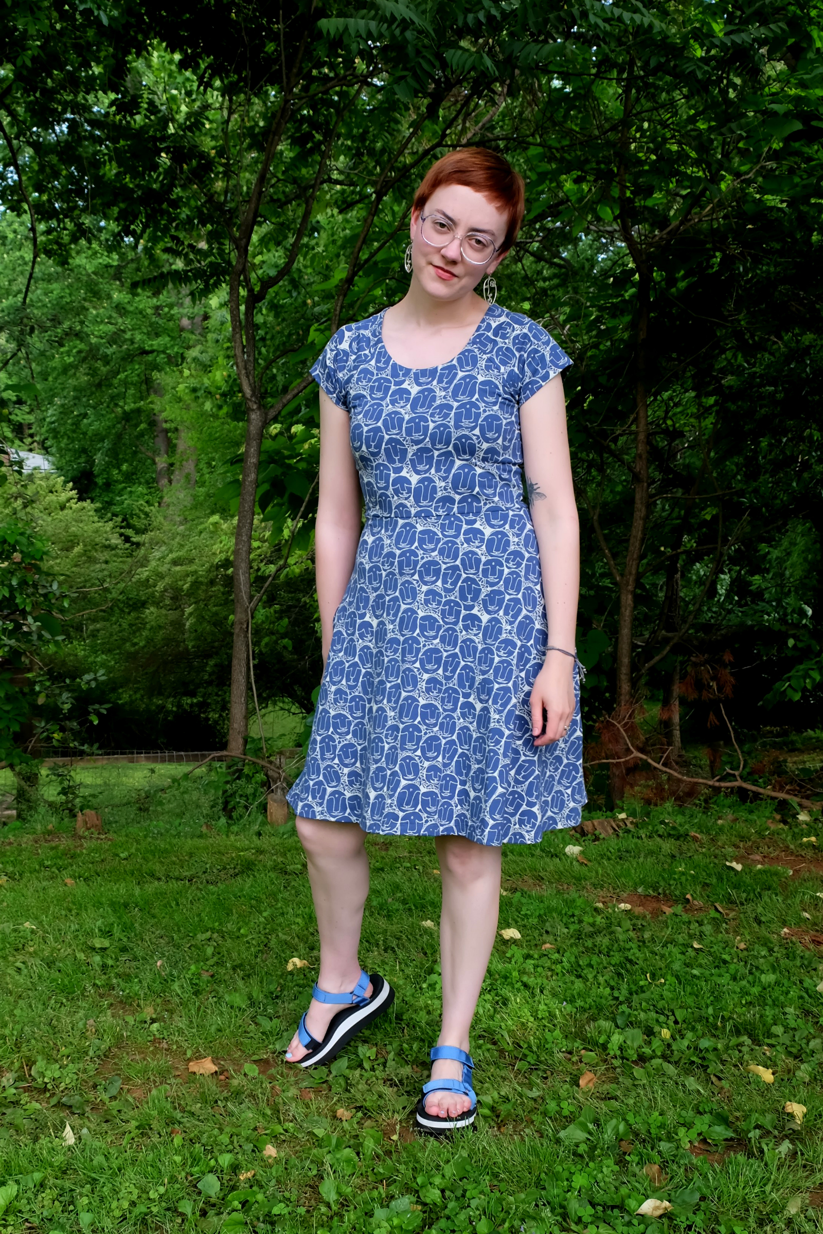 Leah stands in verdant yard wearing a blue dress with faces on it from Mata Traders - Mata Traders Dress