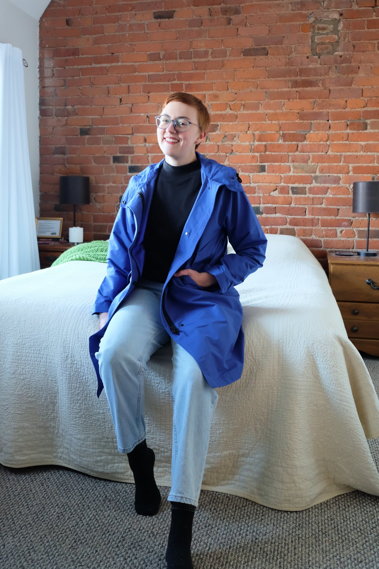 Leah sits on a bed in front of brick wall wearing a blue jacket - Everlane ReNew Anorak Review