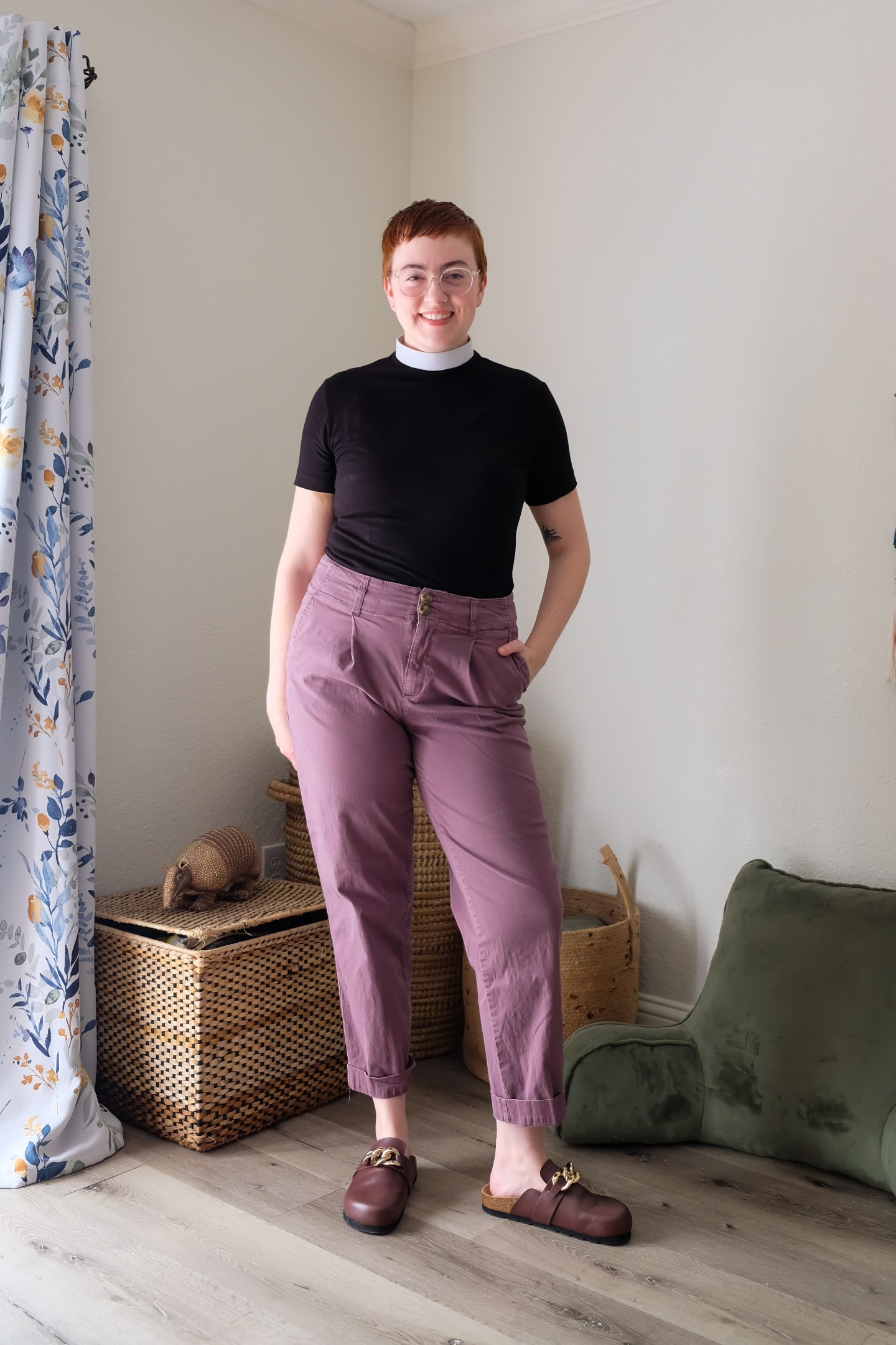 Leah stands in bedroom wearing a black clergy shirt with white collar, purple pants, and burgundy clogs - Alohas Sustainable Clogs Review