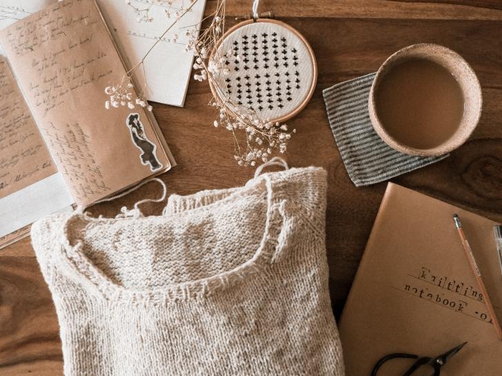 flat lay of cream sweater on wood table with coffee cup, scissors, and journal