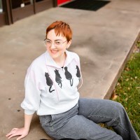 Leah sits on curb wearing sweatshirt with cats on it and gray pants