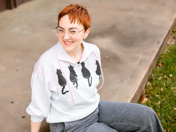 Leah sits on curb wearing sweatshirt with cats on it and gray pants